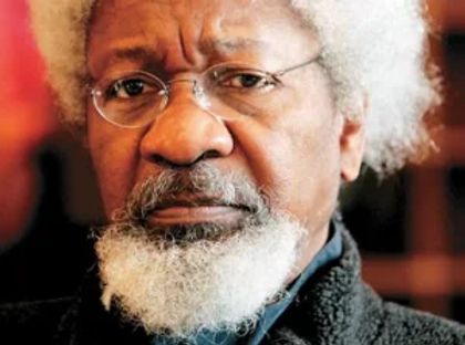 Elderly man with white hair and beard, wearing glasses and a dark jacket, looks serious. Soft focus background with warm tones.