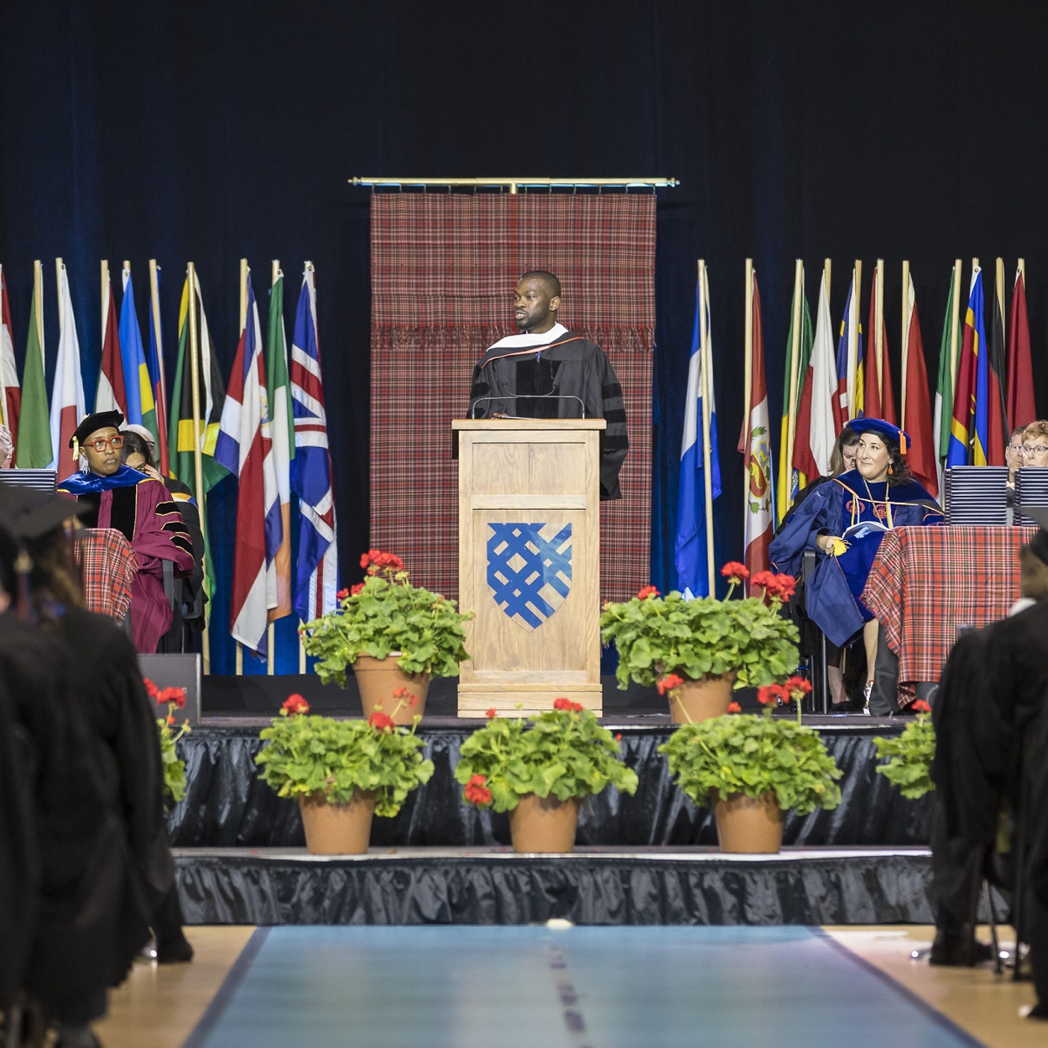 Graduation ceremony with a speaker at podium, flags in the background, and faculty in academic robes seated nearby; flowers decorate the stage.