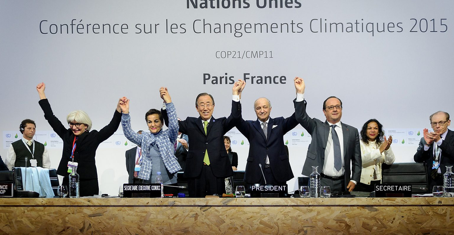 Diverse group celebrates with raised hands at COP21, Paris. Conference backdrop, joyful mood, formal attire, delegates clapping.