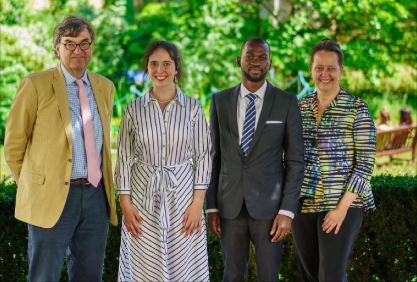 Four people pose smiling in a lush green garden. They wear formal and casual attire in varying colors. Bright and sunny atmosphere.