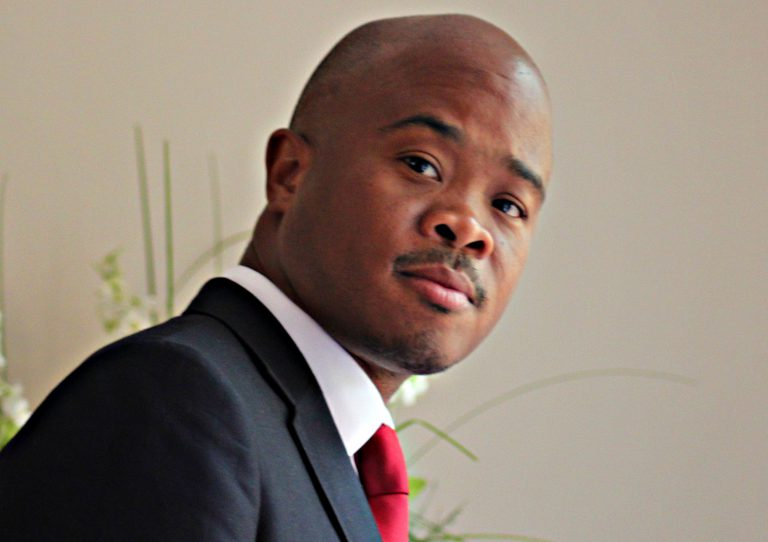 Man in black suit with red tie looks towards the camera, standing against a light background with green plant details. Mood is contemplative.