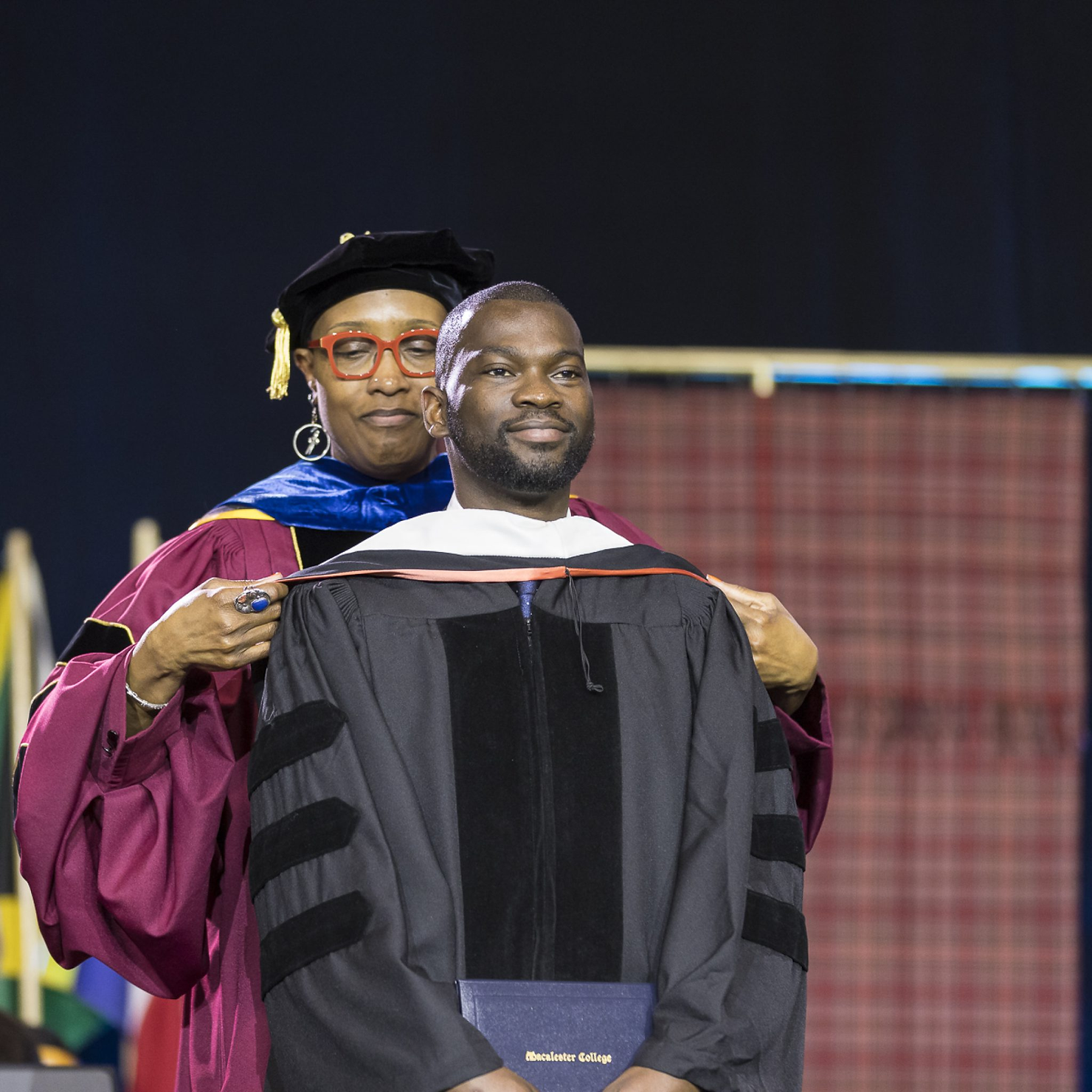 A person in academic attire places a hood on a graduate holding a diploma at a ceremony. Flags are in the background. The mood is celebratory.