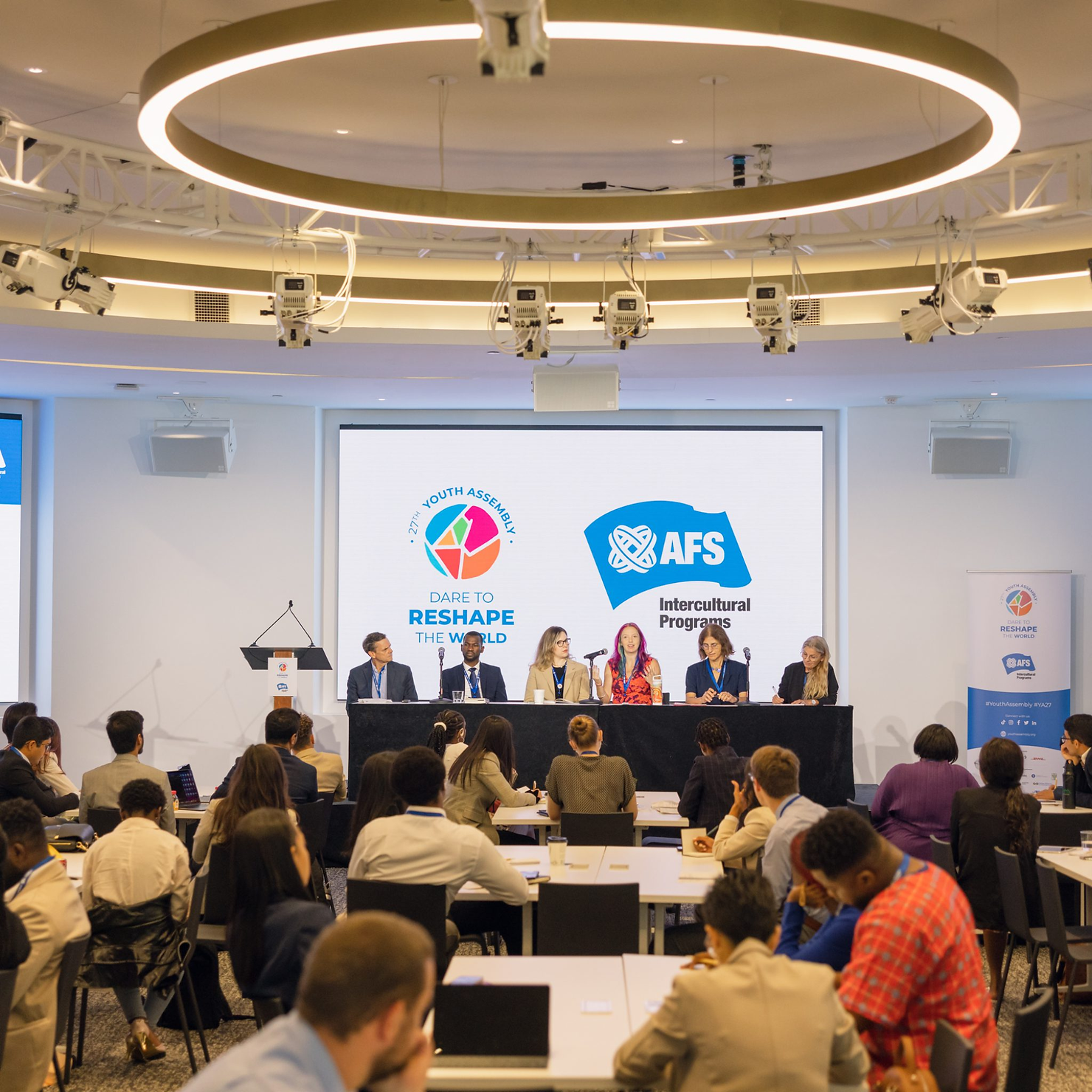 Panel of six people speaking on a stage with a screen displaying "AFS Intercultural Programs" and "Dare to Reshape the World" logo. Audience in foreground.