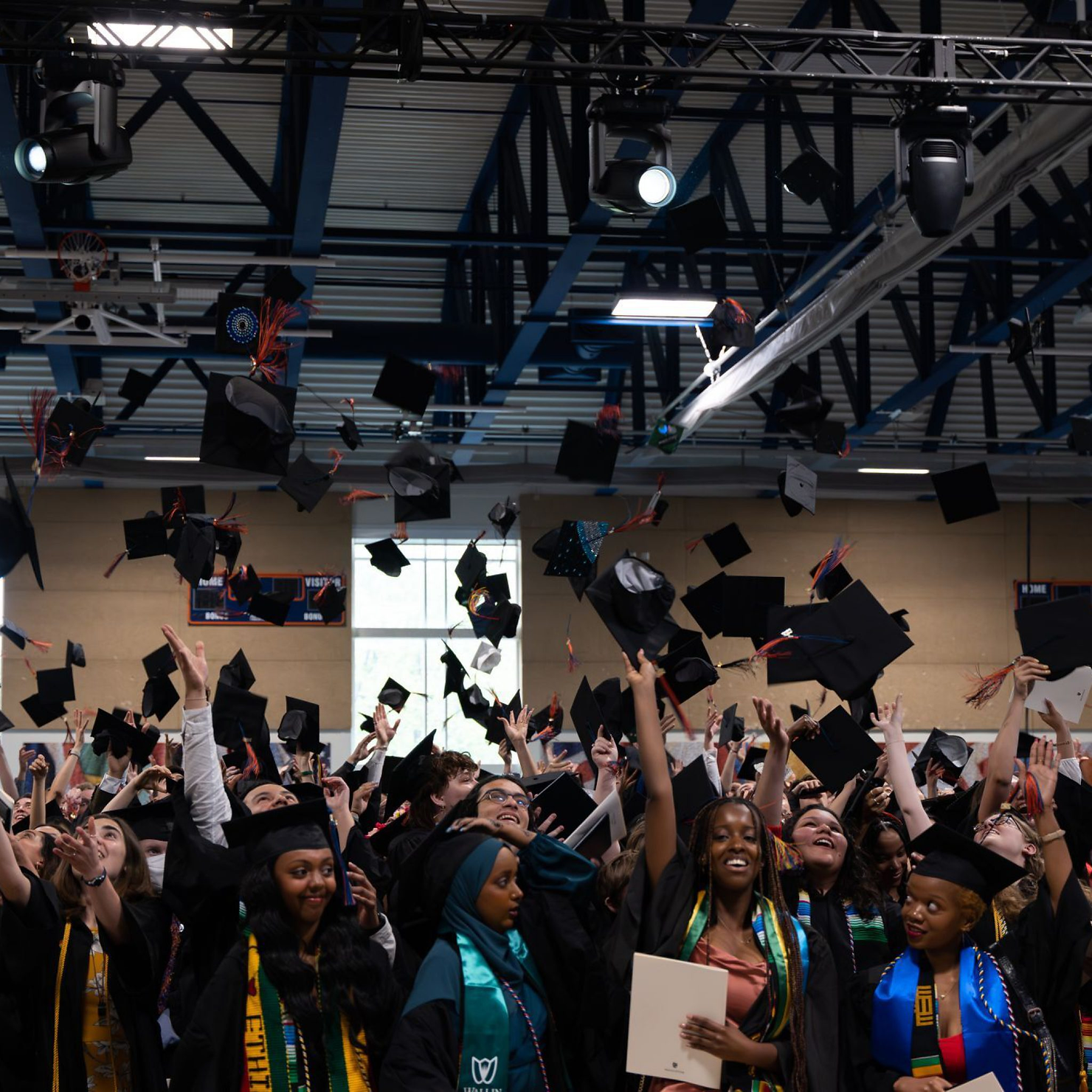 Graduates joyfully toss caps in a gymnasium, celebrating their achievement. The scene is lively with black caps mid-air and vibrant sashes.