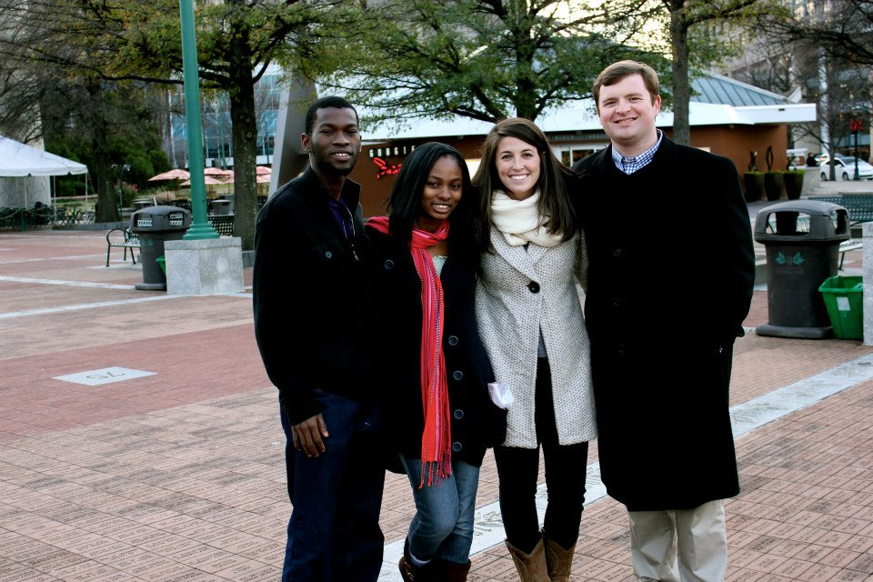 Four people smiling in a park on a chilly day, wearing coats. Trees and a kiosk are in the background. The mood is cheerful and relaxed.