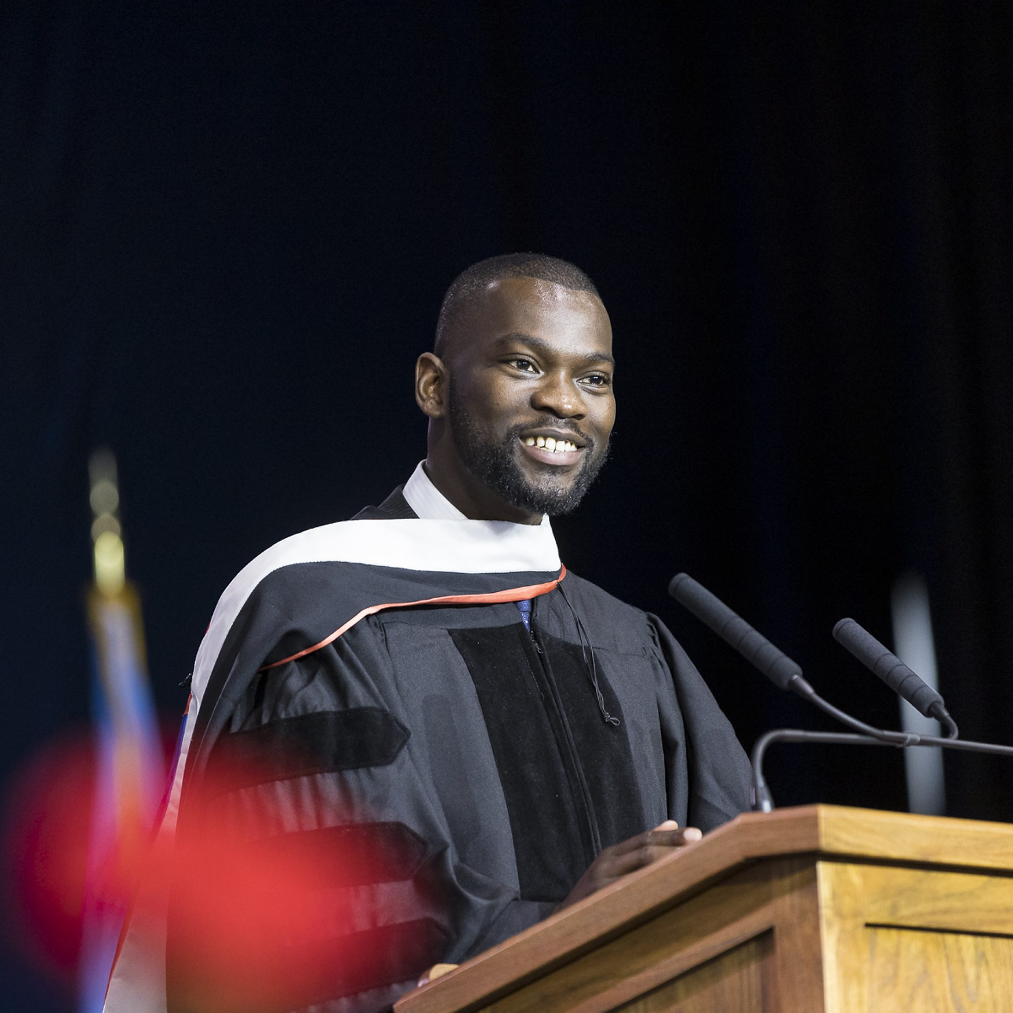 Man in academic regalia smiling, speaking at a podium with microphones. Dark background with a blurred flag visible. Formal and positive mood.